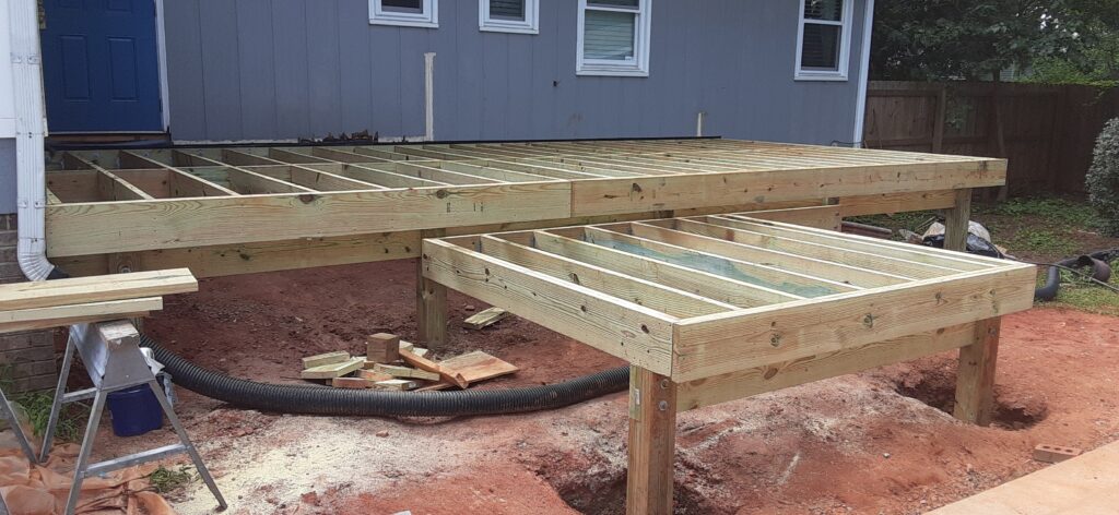 The framework of a new wooden deck under construction in the backyard of a blue house. The structure of the deck is partially complete with visible support beams and joists.