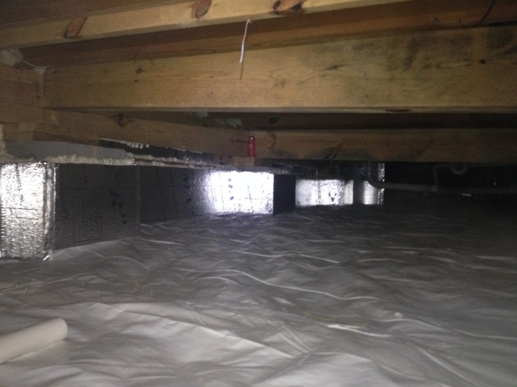 Underneath a house, a crawl space is shown with a new vapor barrier installed on the ground. The reflective insulation on the walls and the wooden beams of the house's substructure are visible, indicating measures taken for moisture control and energy efficiency.