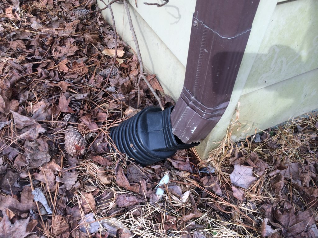 A close-up view of a brown downspout improperly connected to a black flexible adapter intended to direct water away from the foundation of a house.