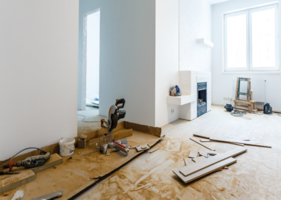 A spacious room undergoing renovation with protective paper on the floor, a miter saw and various construction tools scattered around. The room features a modern fireplace and large windows