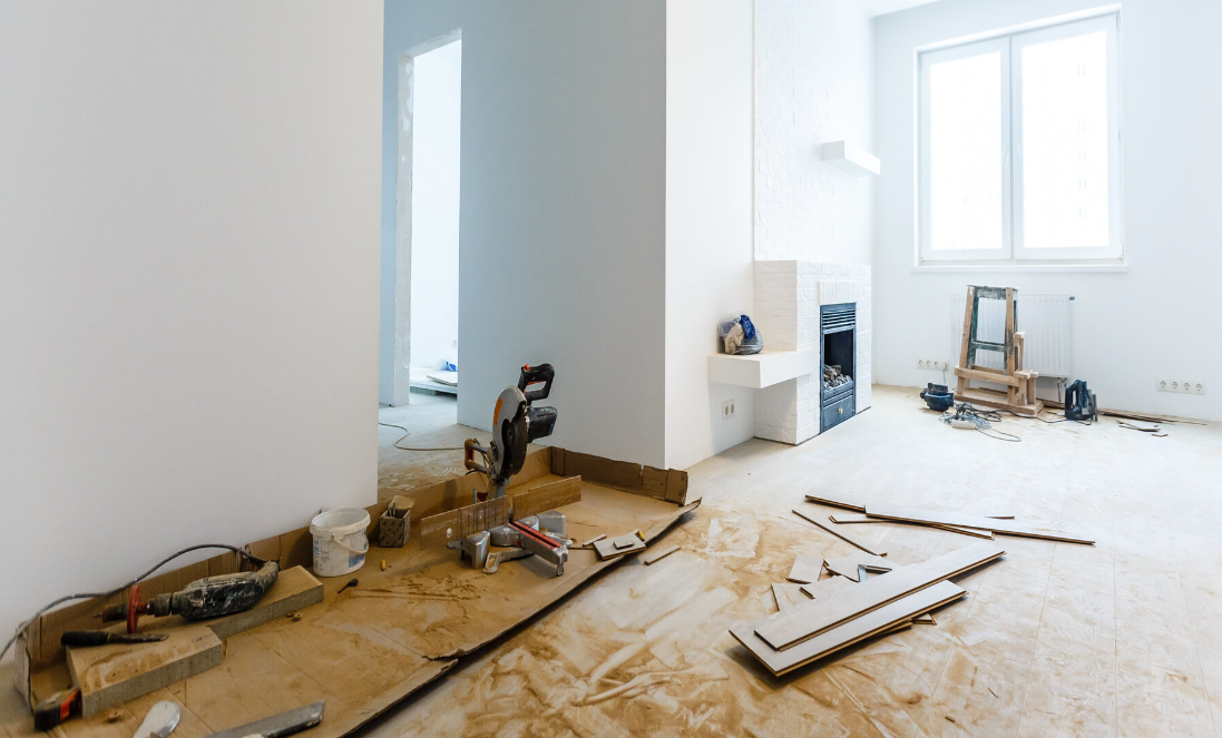 A spacious room undergoing renovation with protective paper on the floor, a miter saw and various construction tools scattered around. The room features a modern fireplace and large windows