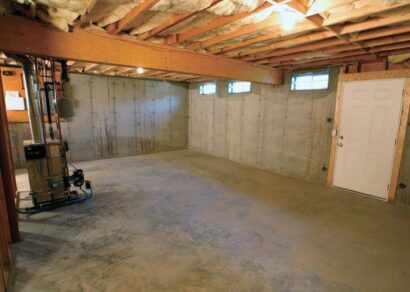 An unfinished basement with exposed insulation in the ceiling and concrete walls. The floor appears bare, and there is a furnace and utility meter installed on the left, with a white door on the right providing entry or exit.
