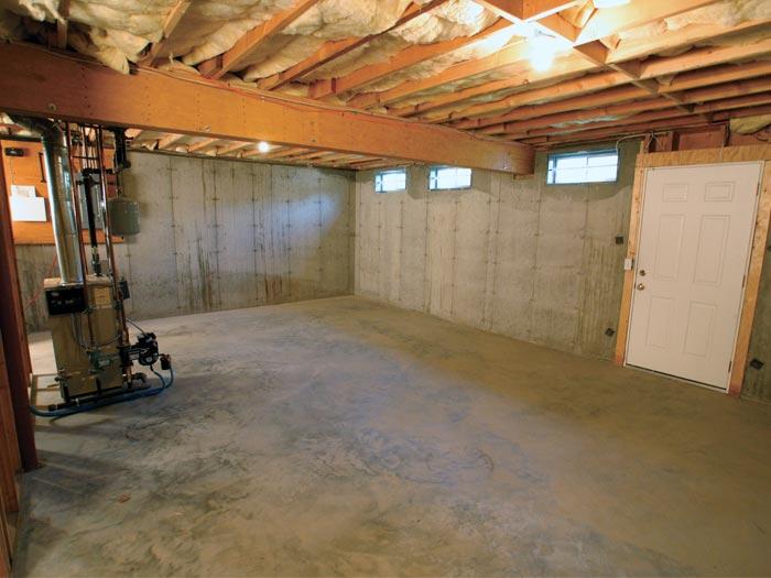 An unfinished basement with exposed insulation in the ceiling and concrete walls. The floor appears bare, and there is a furnace and utility meter installed on the left, with a white door on the right providing entry or exit.