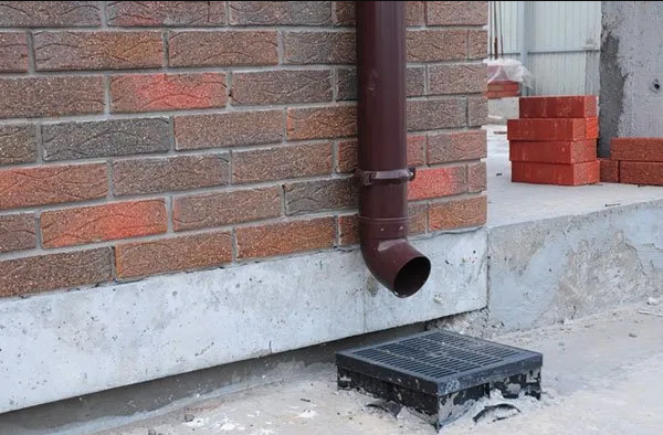 Exterior drainage detail showing a brown downspout directing water into a grate on the ground designed to catch and divert rainwater.