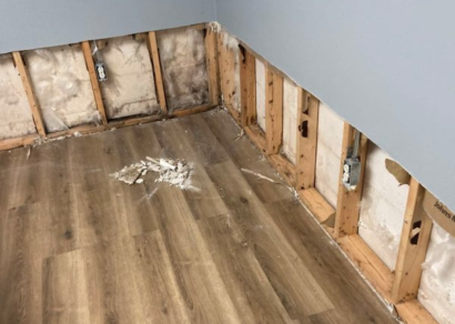 An interior corner of a room showing damage to the lower section of the walls. The drywall has been removed, exposing the insulation and wooden studs. Mold or water damage is visible on the insulation and the wood.