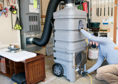 A technician in a garage, kneeling and adjusting a large, gray air duct cleaning machine with a flexible hose attached to the central air system.