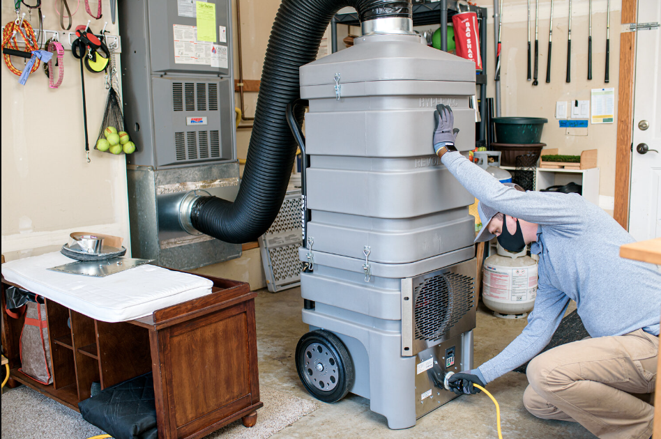 A technician in a garage, kneeling and adjusting a large, gray air duct cleaning machine with a flexible hose attached to the central air system.