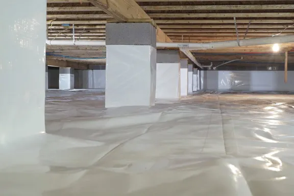 A house crawlspace with a white vapor barrier installed on the ground and wrapped around the support columns.