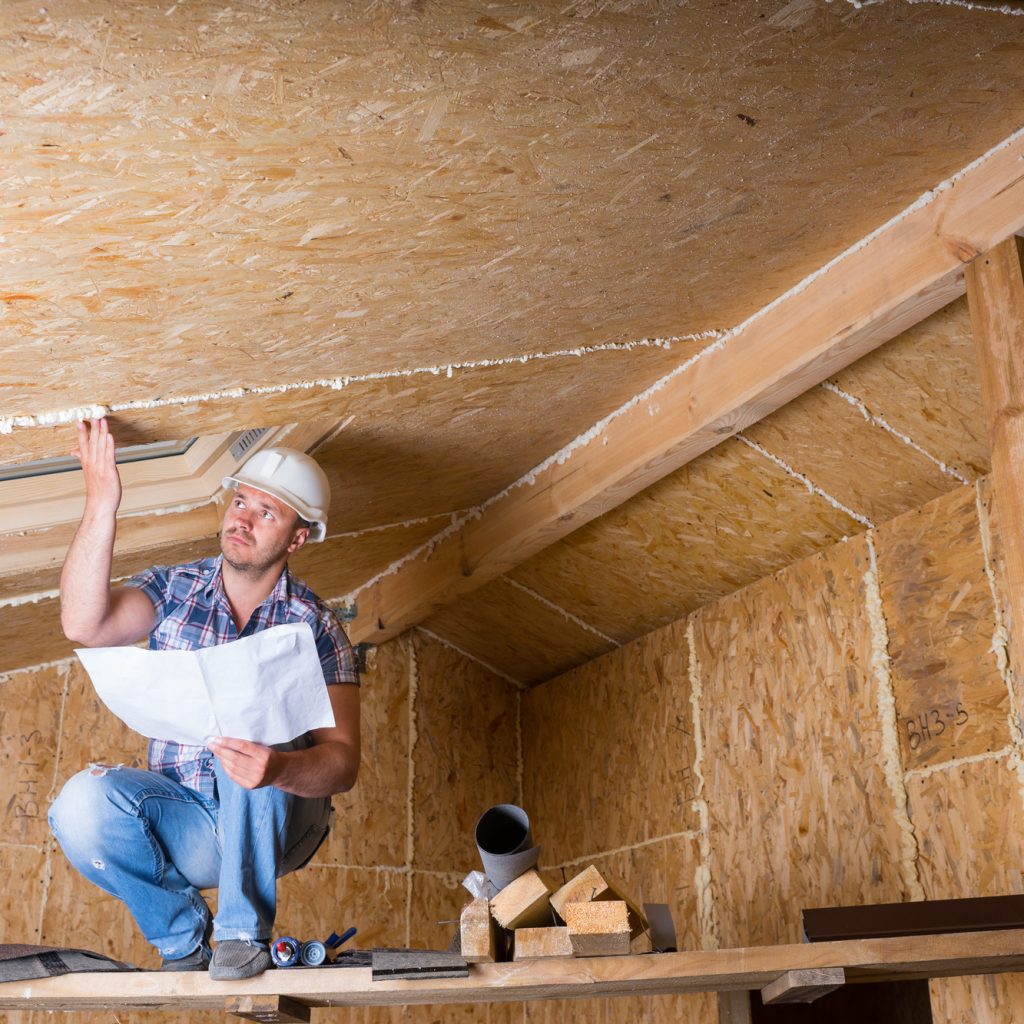 A construction professional crouched in an attic, examining the wooden framework and insulation. He holds plans in one hand, and his expression is focused, suggesting careful assessment of the work to be done. Tools and building materials are within reach, indicating an ongoing installation or inspection process.