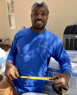 A smiling man wearing a blue shirt with The Solutions Group company logo. He's holding a measuring tape, and wearing a headlamp while standing in front of building insulation materials.